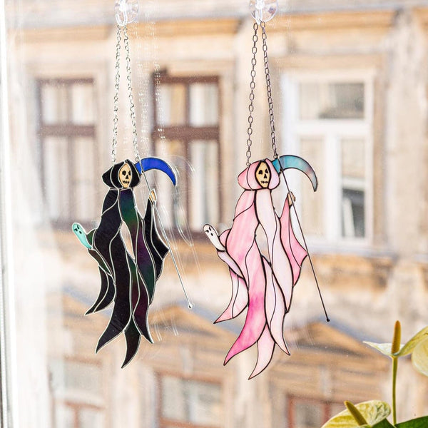 Two hanging stained-glass suncatchers shaped like cloaked figures, one black with blue accents and one pink with teal accents, displayed on a window in front of a blurred building facade and green plant leaves.