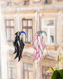 Two hanging stained-glass suncatchers shaped like cloaked figures, one black with blue accents and one pink with teal accents, displayed on a window in front of a blurred building facade and green plant leaves.
