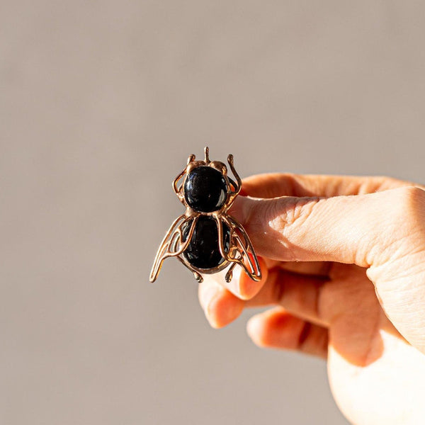 A hand holding a small metallic bee-shaped accessory against a plain warm gray background.