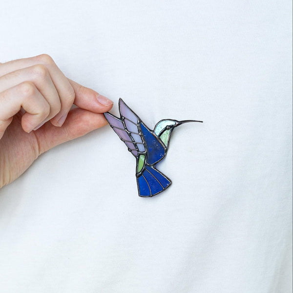 A close-up of a person wearing a white t-shirt and holding a small enamel pin shaped like a hummingbird. The pin features blue, purple, green, and black details.