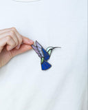 A close-up of a person wearing a white t-shirt and holding a small enamel pin shaped like a hummingbird. The pin features blue, purple, green, and black details.