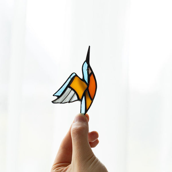 A hand holds a stained-glass-style hummingbird ornament against a bright white curtain background. The bird has light blue, orange, yellow, gray, and black sections outlined in dark metal.