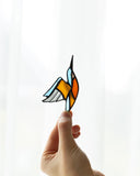 A hand holds a stained-glass-style hummingbird ornament against a bright white curtain background. The bird has light blue, orange, yellow, gray, and black sections outlined in dark metal.