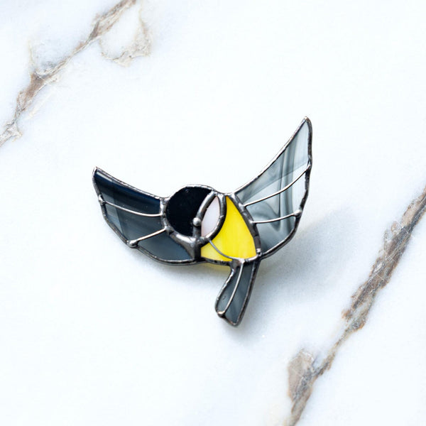 A bird-shaped enamel pin with black wings, a yellow chest, and a black-and-white face, outlined in silver metal, resting on a white marble surface.