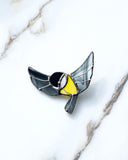 A bird-shaped enamel pin with black wings, a yellow chest, and a black-and-white face, outlined in silver metal, resting on a white marble surface.