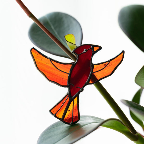 A stained glass bird ornament shaped like a cardinal with orange wings and a red body, hanging near green leaves on a plant stem against a bright white background.