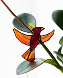 A stained glass bird ornament shaped like a cardinal with orange wings and a red body, hanging near green leaves on a plant stem against a bright white background.