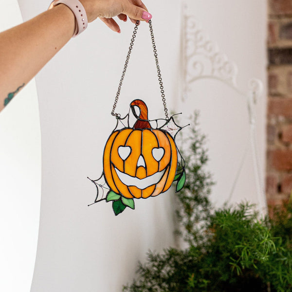 A hand holds a stained glass pumpkin suncatcher with a hanging chain, positioned in front of a white wall with a brick column and potted plants in the background.