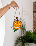 A hand holds a stained glass pumpkin suncatcher with a hanging chain, positioned in front of a white wall with a brick column and potted plants in the background.