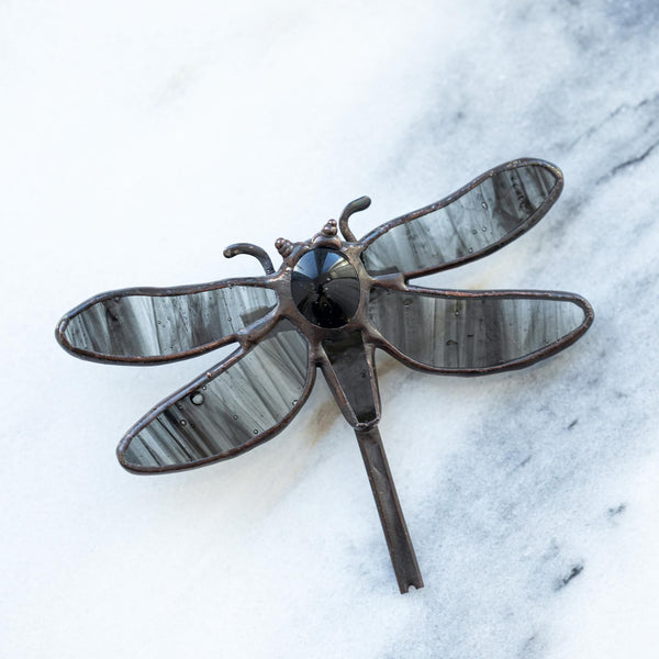 A dragonfly-shaped brooch with dark gray and black marbled wings and a dark metal body, displayed on a white and gray marble surface.