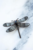 A dragonfly-shaped brooch with dark gray and black marbled wings and a dark metal body, displayed on a white and gray marble surface.